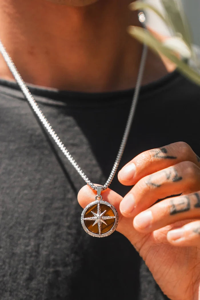 A person in a black shirt displays the Relentless Betrayal Guide Of The Tiger Necklace, showcasing the handcrafted silver pendant with a Tigers Eye stone and star design as sunlight highlights its brown background. Their tattooed hand holds the jewelry.
