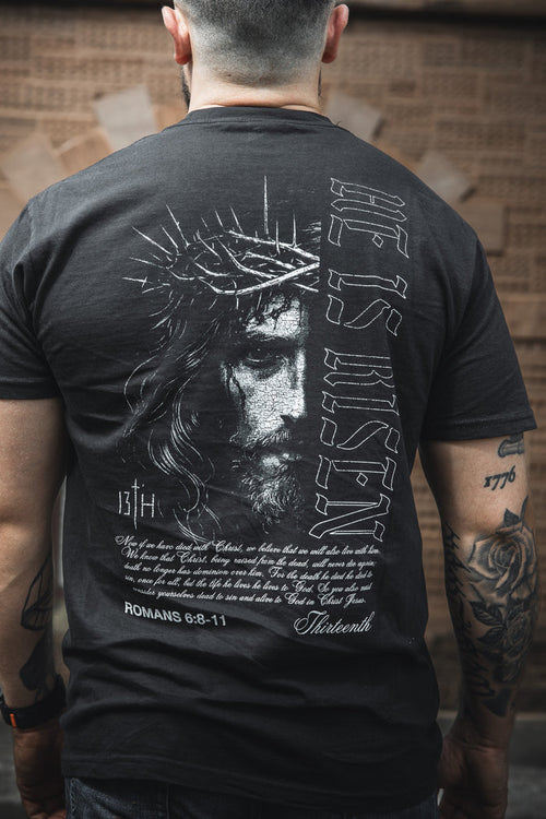 Someone wears the 13th brand "He is Risen Premium Tee," featuring an illustrated Jesus with a crown of thorns, "HE IS RISEN" text, and Romans 6:8-11 on the back—a bold Christian faith shirt.