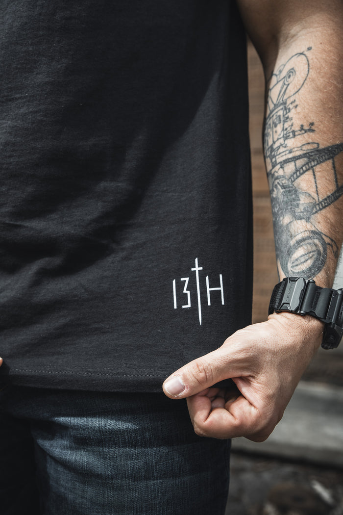 A person wears the He is Risen Premium Tee by 13th, featuring a white 13TH logo near the hem, with blue jeans, a black watch, and a tattoo sleeve—faith apparel for those standing strong in their beliefs.