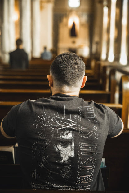 A man with short hair sits alone in a church pew, wearing the 13th He is Risen Premium Tee—a black shirt featuring Jesus crowned with thorns and "HE IS RISEN," boldly expressing his faith through this stylish Christian apparel.