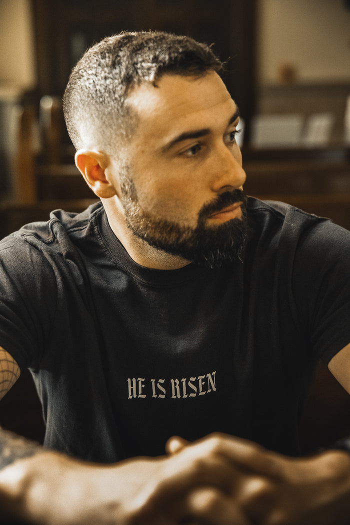 A man with short dark hair and a beard sits indoors, wearing the 13th brand He is Risen Premium Tee, a black Christian t-shirt featuring “HE IS RISEN” in white text, with his arms resting in front and looking to the side.