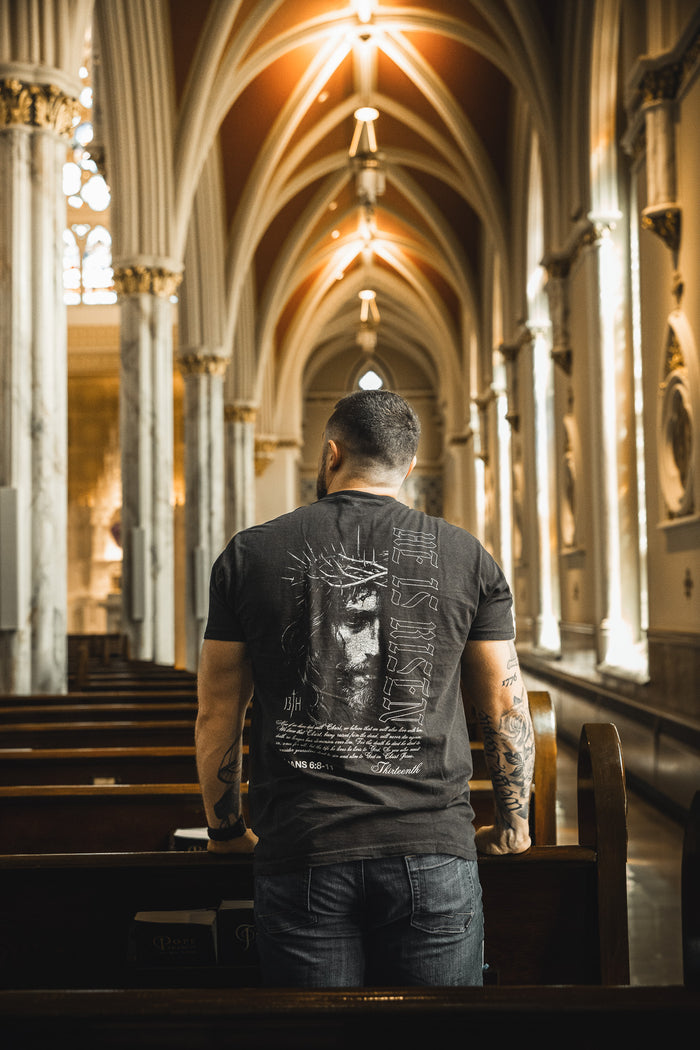 A tattooed man stands in a church pew, wearing the "He is Risen Premium Tee" by 13th. Surrounded by tall arches and stained glass, his bold Christian shirt reflects his faith amid the ornate church interior.