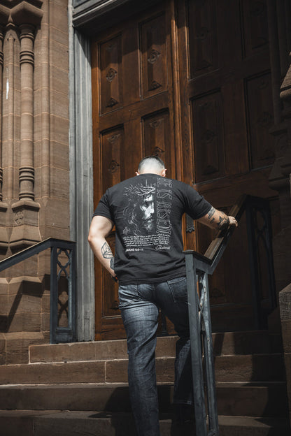 A man in jeans and a black "He is Risen Premium Tee" by 13th walks up stone steps toward a grand wooden door, holding the railing. His faith apparel displays “He is Risen” as ornate stonework frames the scene shot from behind.