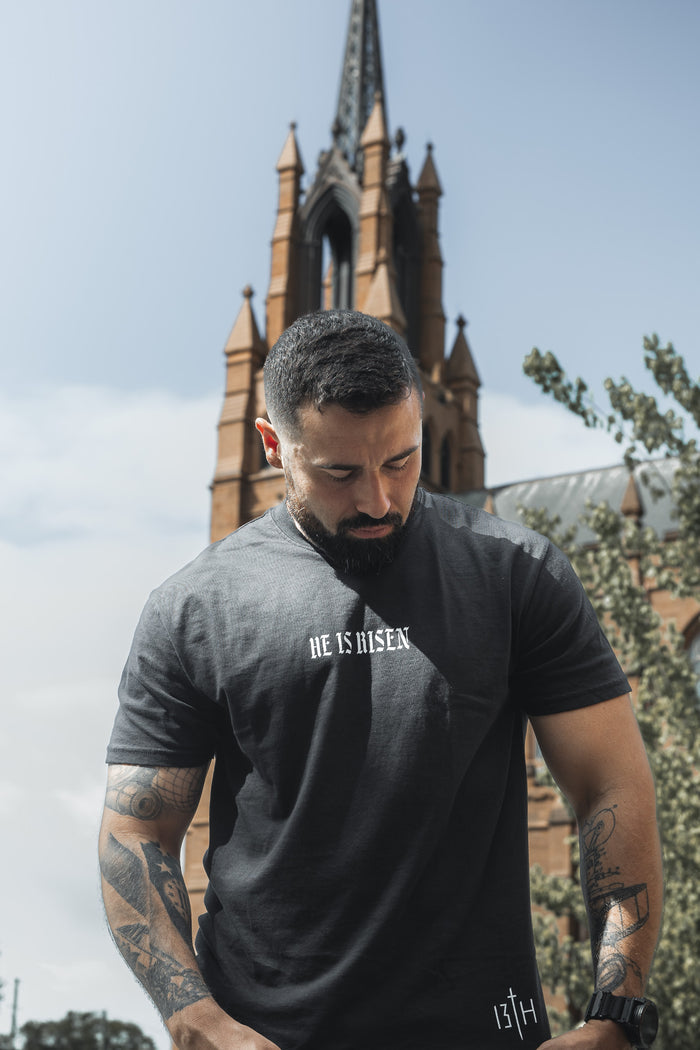 A tattooed man wearing a black "He is Risen Premium Tee" by 13th stands outside a church with a tall spire, looking down calmly. Trees and a partly cloudy sky are in the background.