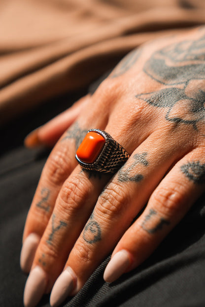 A tattooed hand with long nails displays the Relentless Betrayal Hell Fire Ring, featuring a vivid orange rectangular stone set in a detailed silver band.