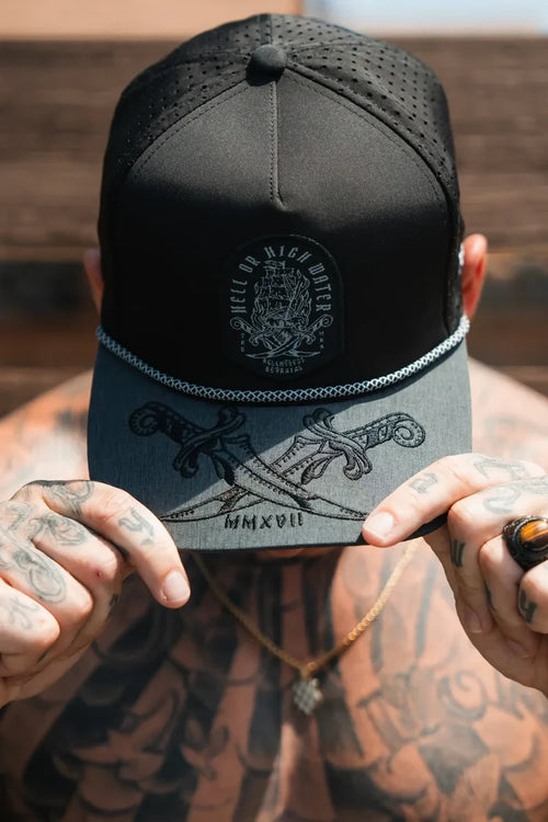 A tattooed person adjusts the brim of a Relentless Betrayal Hell Or High Water Gray Snapback featuring crossed swords and Roman numerals MMXVII, partly hiding their face. They wear a gold necklace and a large stone ring, embodying unyielding resilience.