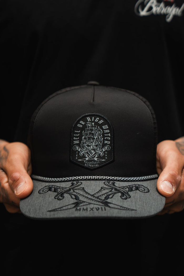 A person holds the Hell or High Water Gray Snapback by Relentless Betrayal, showcasing a decorative patch. The gray brim, adorned with intricate designs and Roman numerals MMXVII, complements their black shirt, partially visible.