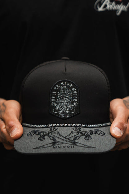 A person holds the Hell or High Water Gray Snapback by Relentless Betrayal, showcasing a decorative patch. The gray brim, adorned with intricate designs and Roman numerals MMXVII, complements their black shirt, partially visible.