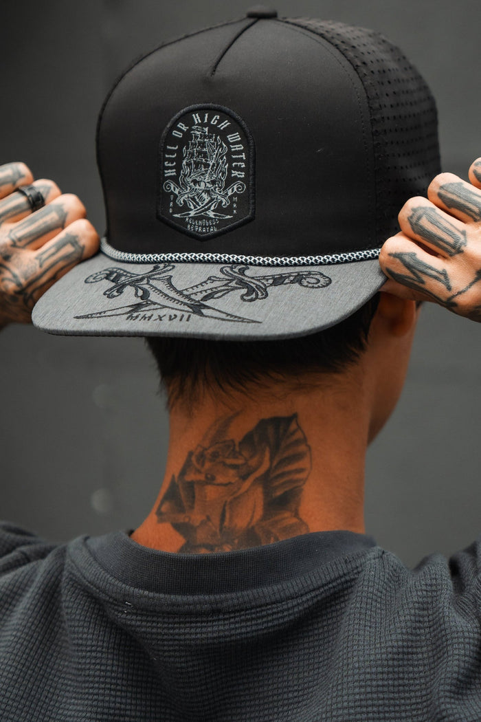 A person wears a gray snapback by Relentless Betrayal, featuring the Hell Or High Water design with crossed swords and ships. Tattoos adorn their neck and hands, as the text HELL OR HIGH WATER boldly emphasizes resilience.
