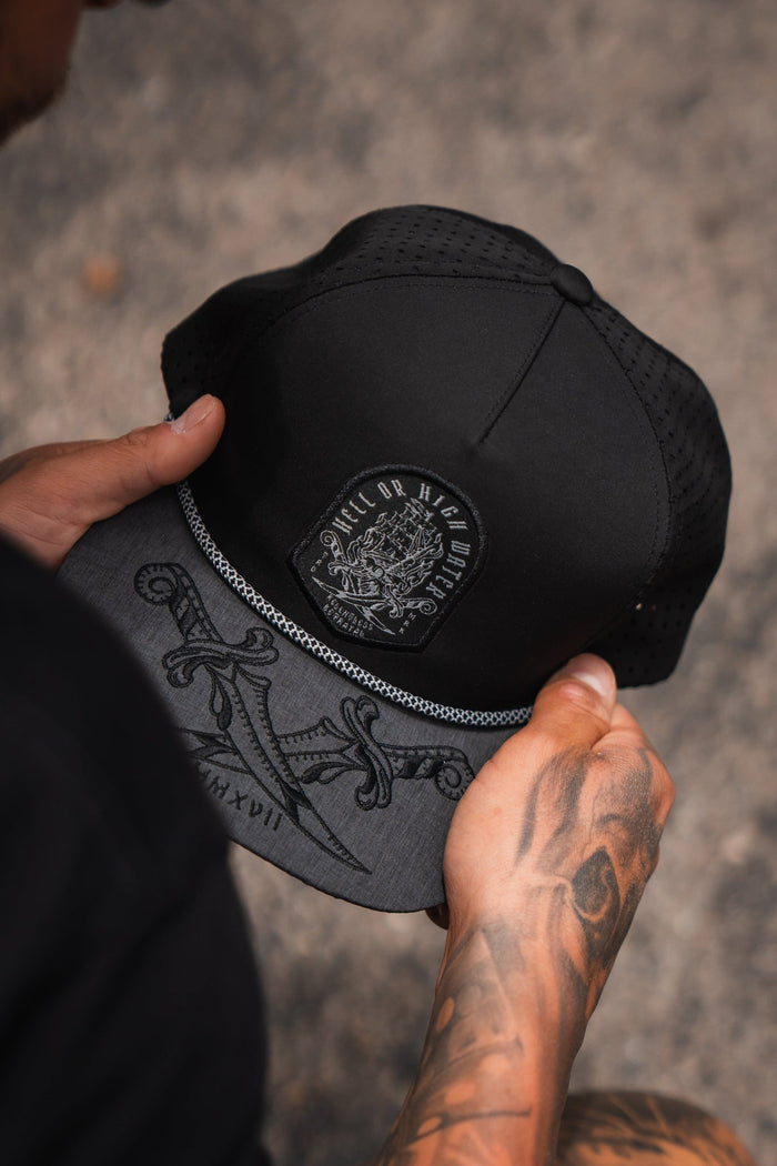 A person with tattooed hands holds the Hell Or High Water Gray Snapback by Relentless Betrayal. This water-resistant hat features an embroidered patch with Kill or Be Killed, a brim with swords, and MMXVII, symbolizing unyielding resilience.