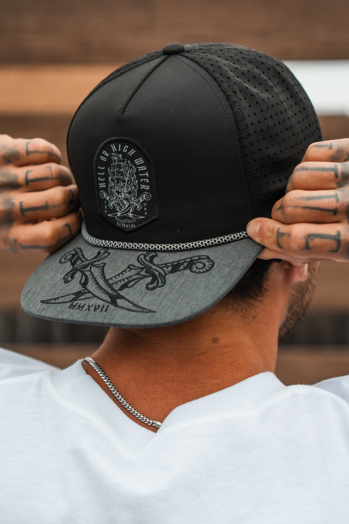 A person wearing the Relentless Betrayal Hell Or High Water Gray Snapback adjusts its fit. The cap, featuring a design patch and artwork on the brim, symbolizes resilience. Tattoos adorn their fingers as they wear a white shirt, with short hair visible.
