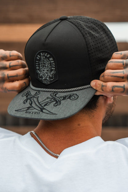 A person wearing the Relentless Betrayal Hell Or High Water Gray Snapback adjusts its fit. The cap, featuring a design patch and artwork on the brim, symbolizes resilience. Tattoos adorn their fingers as they wear a white shirt, with short hair visible.