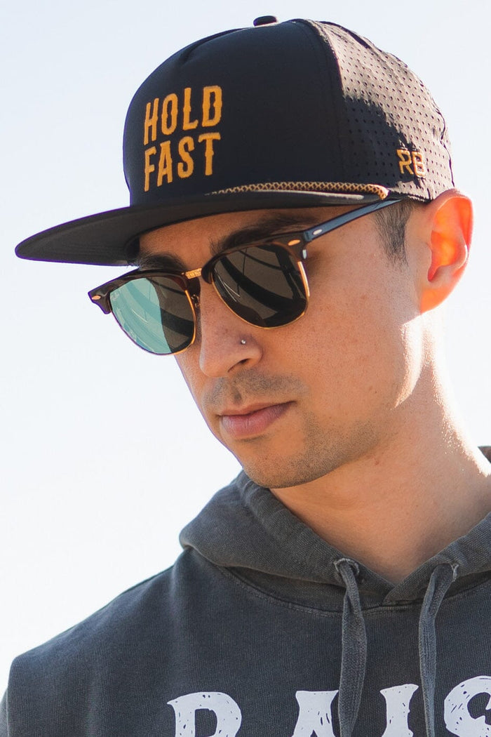 A person wears a Relentless Betrayal Hold Fast Snapback cap with an orange logo, sunglasses, and a grey hoodie. The cap has a flat bill and the individual has a nose piercing, looking downwards against a bright, blurred background.