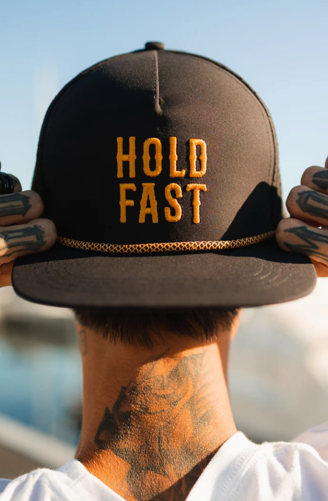 A person is wearing the Hold Fast Snapback by Relentless Betrayal, a black cap adorned with HOLD FAST in yellow and a rope above the flat bill. The handmade, breathable design complements their tattoos as they hold its sides against a blurred background.