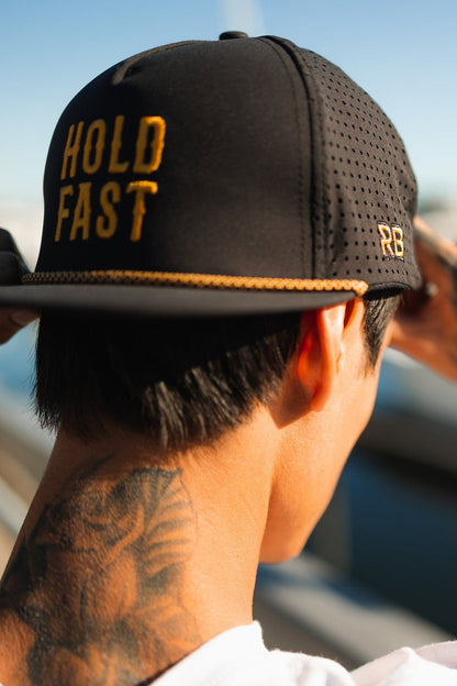 A person adjusts their Relentless Betrayal Hold Fast Snapback, a handmade black cap with yellow embroidery and the words HOLD FAST on the back. The flat bill design complements their lion neck tattoo, set against a blurred outdoor background.