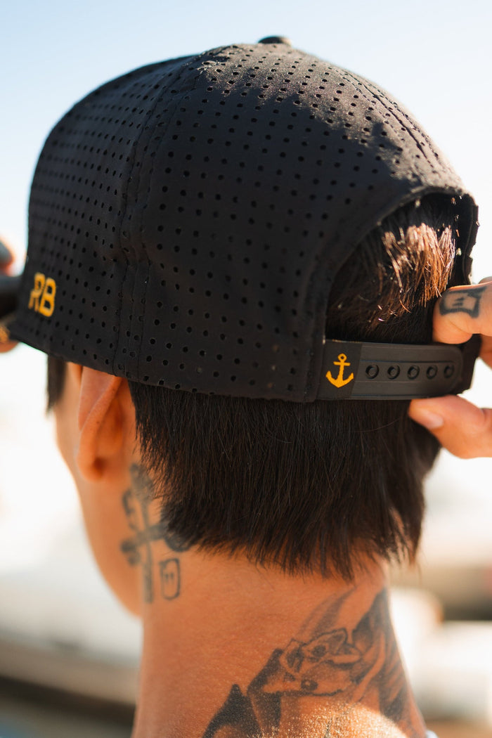 A tattooed person adjusts a Hold Fast Snapback by Relentless Betrayal. The handmade black perforated cap features yellow RB initials and an anchor symbol on the adjustable strap, with a blurred background.