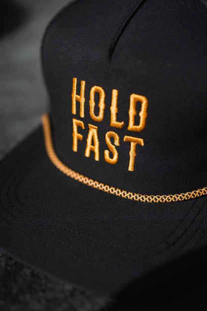 The Hold Fast Snapback by Relentless Betrayal is a black flat bill cap with HOLD FAST in bold yellow embroidery on the front, decorative yellow stitching on the brim, and a breathable snapback design for comfort.