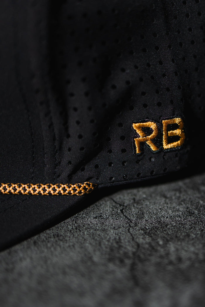 A close-up of the black perforated fabric from the handmade Hold Fast Snapback by Relentless Betrayal shows gold-stitched RB initials and a decorative stitch pattern, with a textured gray background enhancing the design contrast.