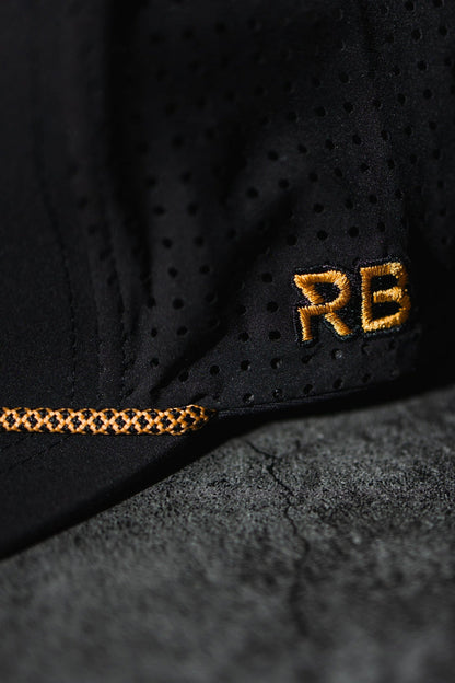 A close-up of the black perforated fabric from the handmade Hold Fast Snapback by Relentless Betrayal shows gold-stitched RB initials and a decorative stitch pattern, with a textured gray background enhancing the design contrast.