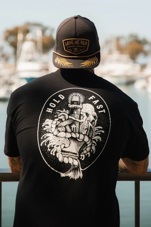 A person in a black Relentless Betrayal Lost at Sea cap and a Hold Fast T-Shirt—Black leans on a railing, gazing over a marina with blurred boats and water in the background.