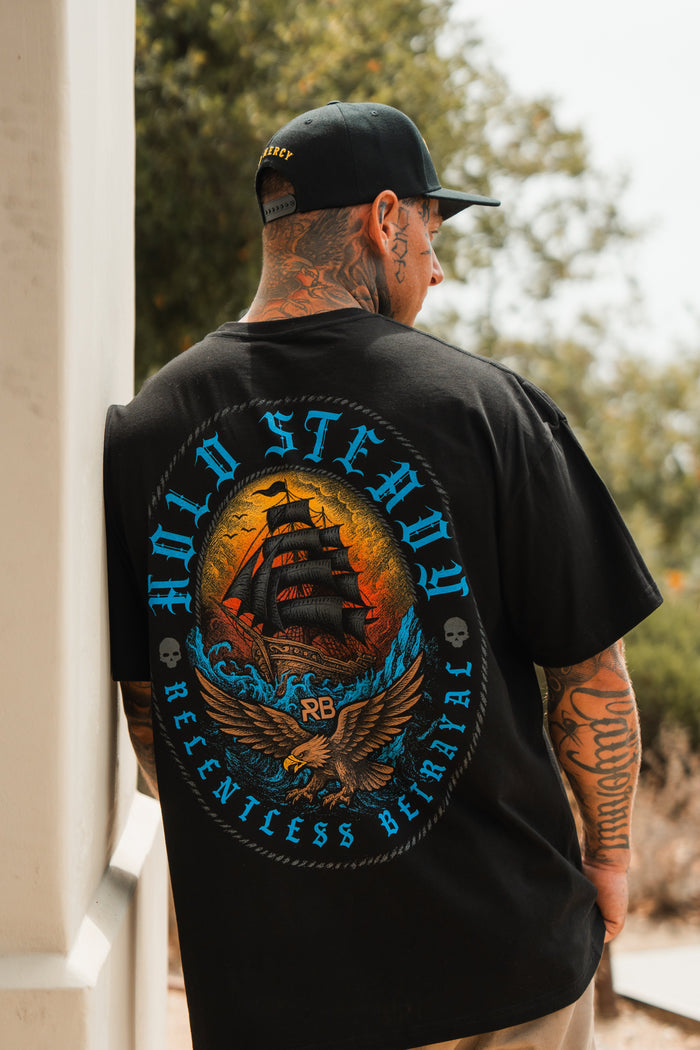 A tattooed person outdoors wears the Relentless Betrayal Hold Steady Blue Premium Oversized Tee, a black shirt featuring a ship and eagle graphic with bold blue and orange text.