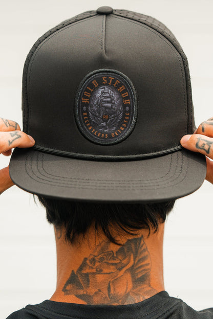 A tattooed individual adjusts a Relentless Betrayal Hold Steady Ghost Snapback, featuring a skull and text patch. Their face is hidden as the photo is taken from behind.