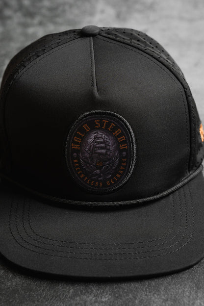 A close-up of the Hold Steady Ghost Snapback by Relentless Betrayal—a premium snapback baseball cap with a round patch reading HOLD STEADY BREATHLESS BETRAYED in orange and showcasing a coiled snake.