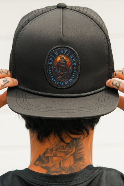 A person adjusts the brim of the Relentless Betrayal Hold Steady Snapback, featuring a circular patch and water-resistant design. Visible finger tattoos and a large floral neck tattoo stand out against a plain, light-colored background.