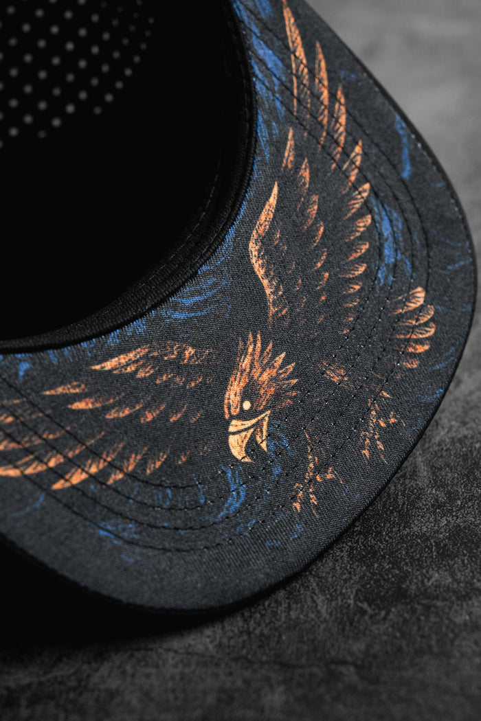Close-up of the Relentless Betrayal Hold Steady Snapback, a premium black hat with an illustrated eagle in orange and blue on the brim, displayed on a textured dark gray surface.