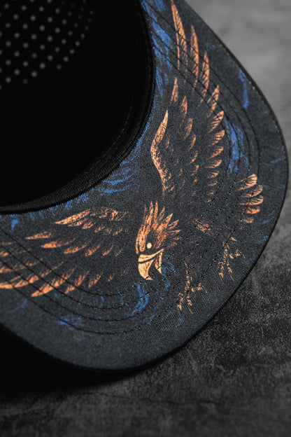 Close-up of the Relentless Betrayal Hold Steady Snapback, a premium black hat with an illustrated eagle in orange and blue on the brim, displayed on a textured dark gray surface.