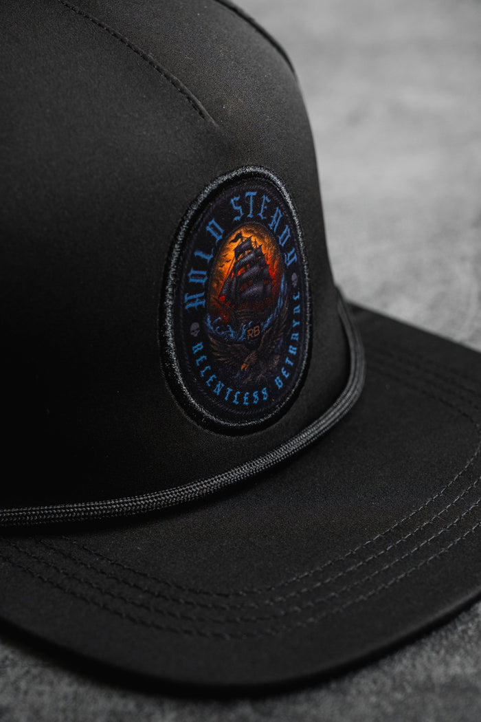 A close-up of the Hold Steady Snapback by Relentless Betrayal, featuring a round embroidered ship patch with "HOLD STEADY" and "RELENTLESS BELIEFING." This water-resistant hat rests on a gray surface.