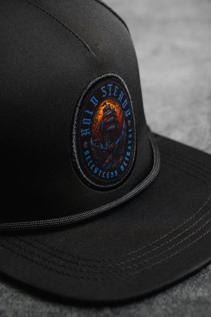 A close-up of the Hold Steady Snapback by Relentless Betrayal, featuring a round embroidered ship patch with "HOLD STEADY" and "RELENTLESS BELIEFING." This water-resistant hat rests on a gray surface.