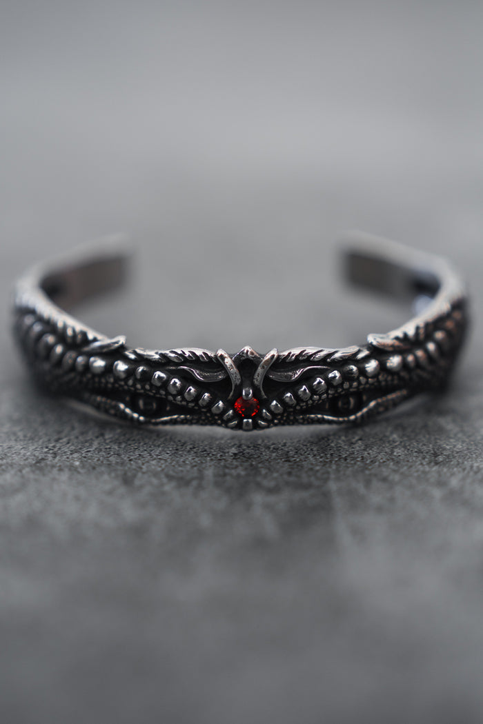 A close-up of the intricately designed Relentless Betrayal Holy Grail Bracelet showcases its dark stainless steel with a dragon motif, detailed scales, wings, and a central red gem. The softly blurred gray background enhances its mythical artifact allure.