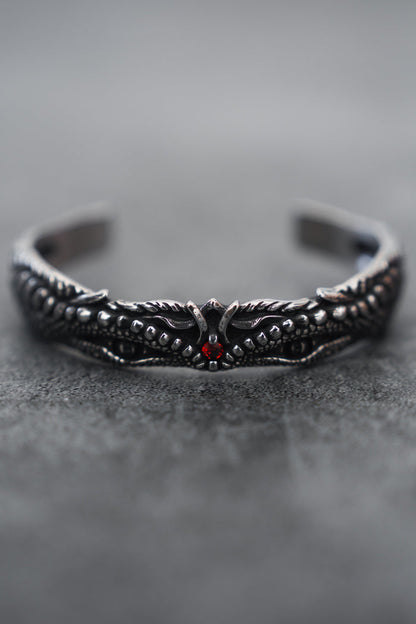 A close-up of the intricately designed Relentless Betrayal Holy Grail Bracelet showcases its dark stainless steel with a dragon motif, detailed scales, wings, and a central red gem. The softly blurred gray background enhances its mythical artifact allure.