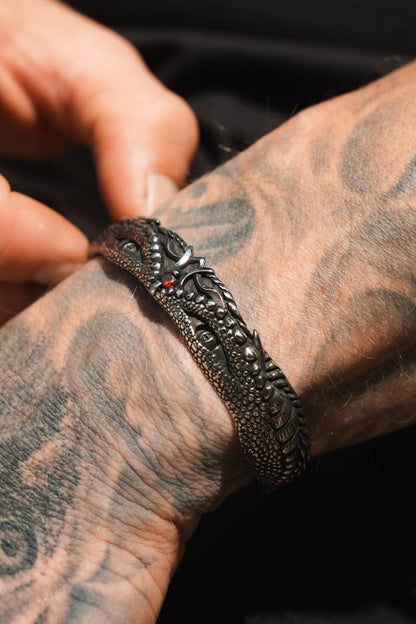 A tattooed hand adjusts Relentless Betrayals Holy Grail Bracelet, a stainless steel masterpiece with intricate designs and red accents, while abstract patterns decorate the skin.