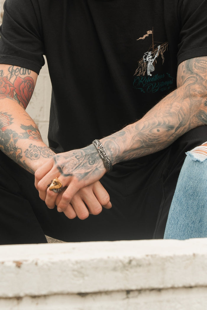 A person with tattooed arms sits wearing a black T-shirt and ripped jeans. Among their bracelets, the Relentless Betrayal Holy Grail Bracelet stands out. A ring adorns their fingers, but their face remains hidden from view.