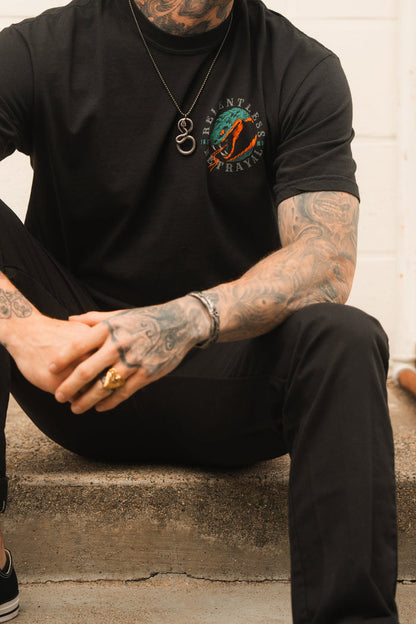 A tattooed person sits on concrete steps, wearing black pants and a t-shirt with a colorful design. Adorned with Relentless Betrayals Holy Grail bracelet and various rings, their hands are clasped. A necklace featuring a ring pendant drapes over the shirt.