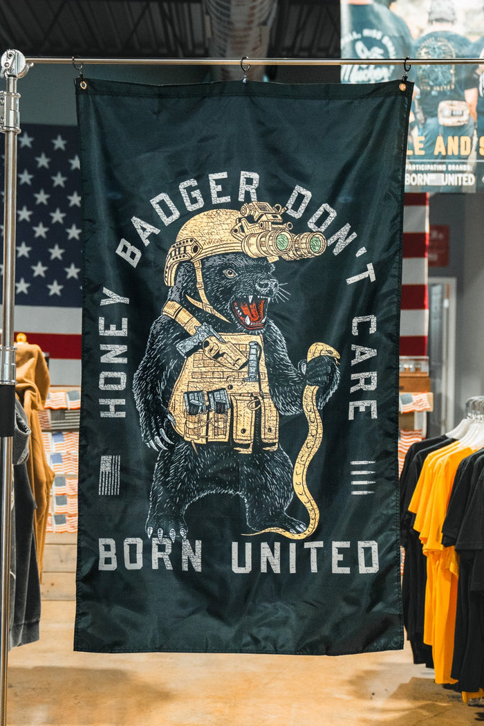 A Honey Badgerin' Flag from Born United Private Label hangs in a clothing store, showing an armored badger with gold gear and the text "HONEY BADGER DON’T CARE" and "BORN UNITED." T-shirts and American flags are seen behind it.