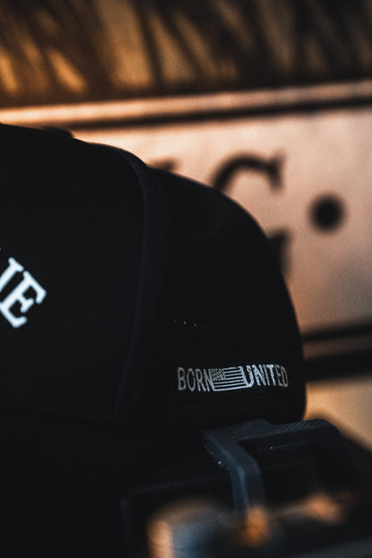 A close-up of the I Am Charlie Black Snapback from Born United Private Label, featuring "BORN UNITED" text and an American flag stitched on the side, set against a blurred background with warm lighting.