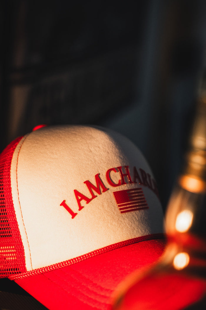 The Born United Private Label “I Am Charlie Red Trucker Snapback” features red and white panels, a curved brim, “I AM CHARL” text, and a stylized American flag on the front—adding bold style to any handmade hat collection.