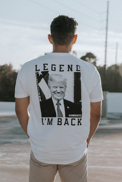 A man stands outside in a Born United Private Label Im Back - #47 - White T-shirt, celebrating a presidents influence. A fence and trees bask under the clear sky in the background.