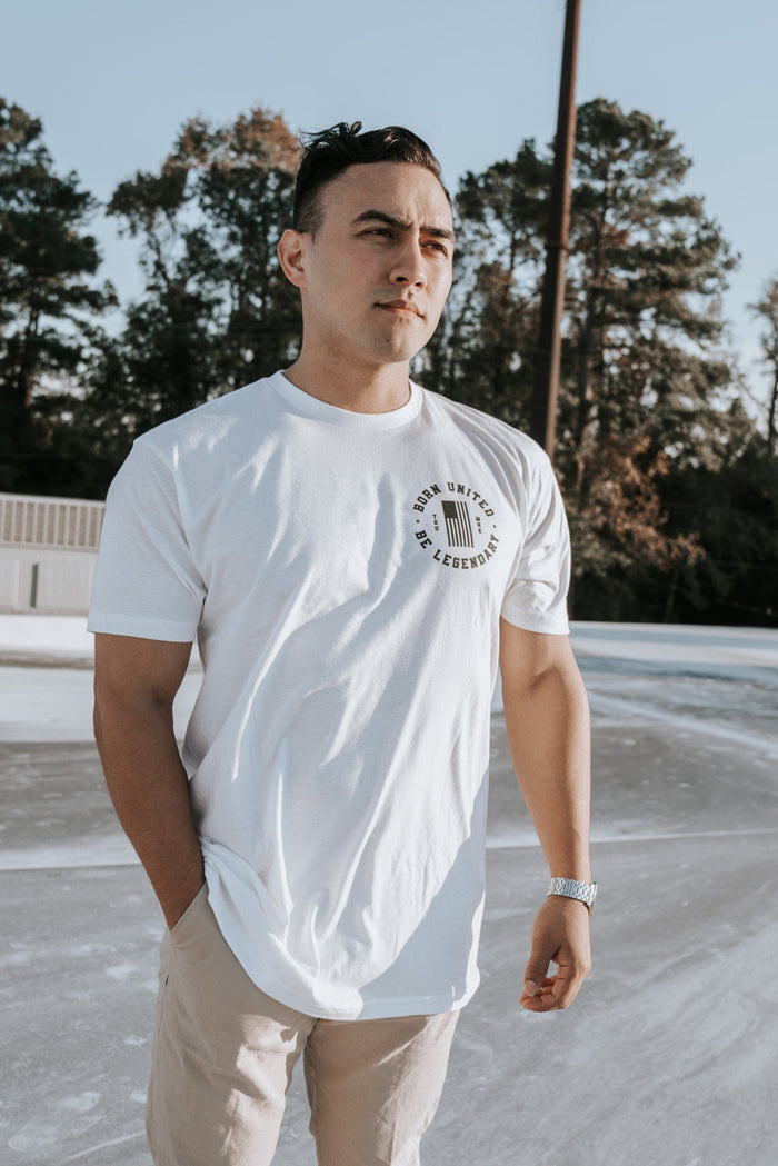 Wearing the Im Back - #47 - White tee by Born United Private Label and beige pants, a man stands outdoors, hand in pocket, gazing into the distance. Trees and utility boxes form the backdrop as he reflects on potential future victories, like a Trump 2024 win.