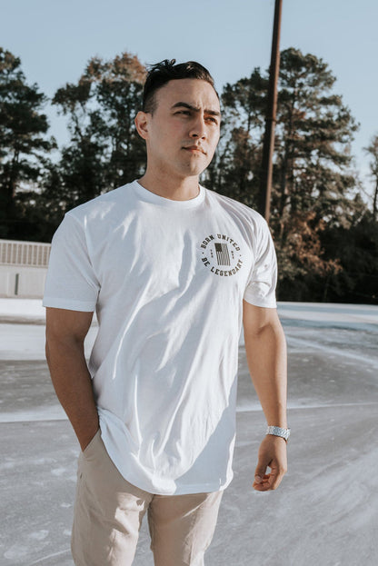 Wearing the Im Back - #47 - White tee by Born United Private Label and beige pants, a man stands outdoors, hand in pocket, gazing into the distance. Trees and utility boxes form the backdrop as he reflects on potential future victories, like a Trump 2024 win.