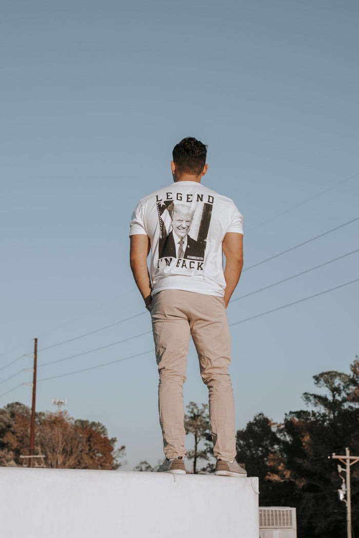 On the edge of a white rooftop, someone wears the Im Back - #47 - White tee by Born United Private Label, emblazoned with LEGEND and a faded graphic, paired with beige pants. The clear blue sky meets trees and power lines in the background, setting an anticipatory scene.