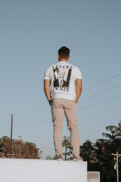 On the edge of a white rooftop, someone wears the Im Back - #47 - White tee by Born United Private Label, emblazoned with LEGEND and a faded graphic, paired with beige pants. The clear blue sky meets trees and power lines in the background, setting an anticipatory scene.