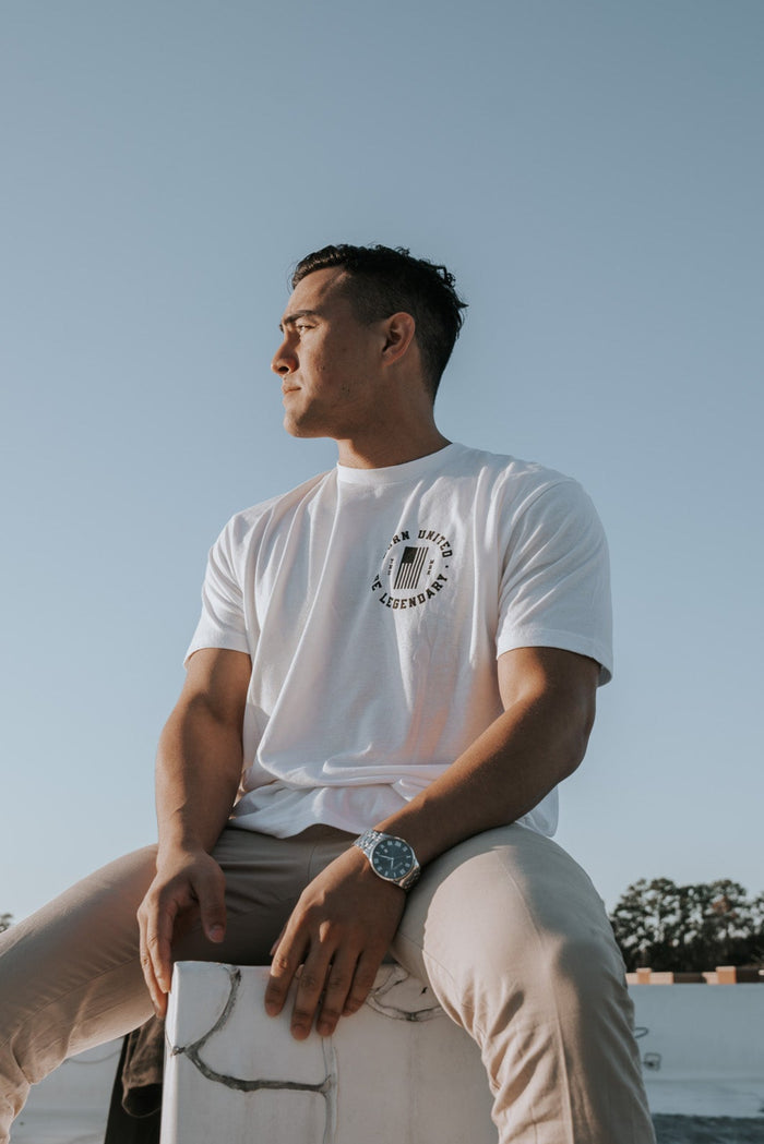 A man wearing the Im Back - #47 - White tee from Born United Private Label and beige pants gazes sideways against a clear blue sky. Sporting a watch, distant trees and rooftops bask in soft daylight, possibly reflecting on the implications of a Trump 2024 win.