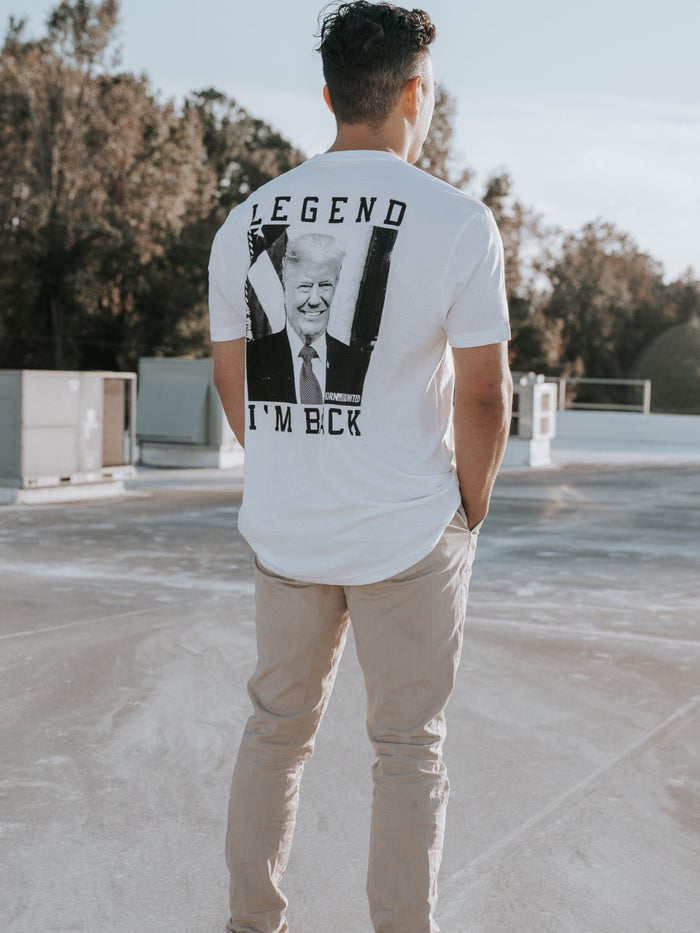 A man stands on a rooftop wearing the Im Back - #47 - White T-shirt from Born United Private Label, hinting at Trumps 2024 win. With hands in pockets, casual pants, and sneakers, he gazes into the clear sky with trees in the background.
