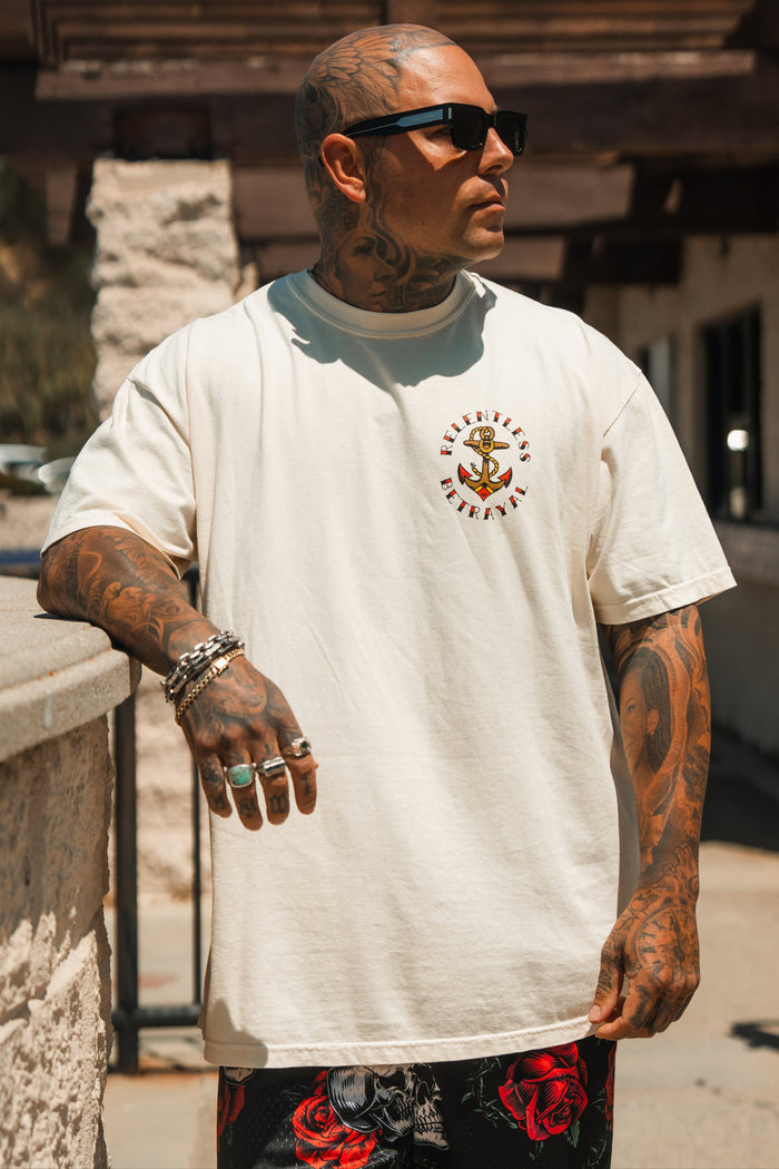 A tattooed man in sunglasses wears the Relentless Betrayal Into the Storm Premium Ivory T-Shirt and black shorts with red roses, standing outdoors by a stone wall and looking to the side.