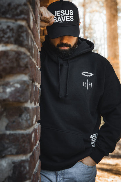 A man in a 13th Jesus Saves Black Snapback and black hoodie with white text stands leaning against a brick wall outdoors, looking down with one hand on the wall.