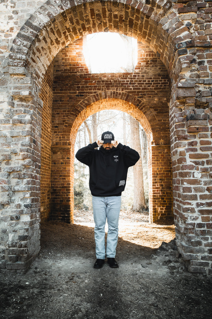 A person wearing a black hoodie, light blue jeans, and a 13th Jesus Saves Black Snapback stands under a brick archway, holding the brim of their cap with both hands as sunlight streams through an opening above.